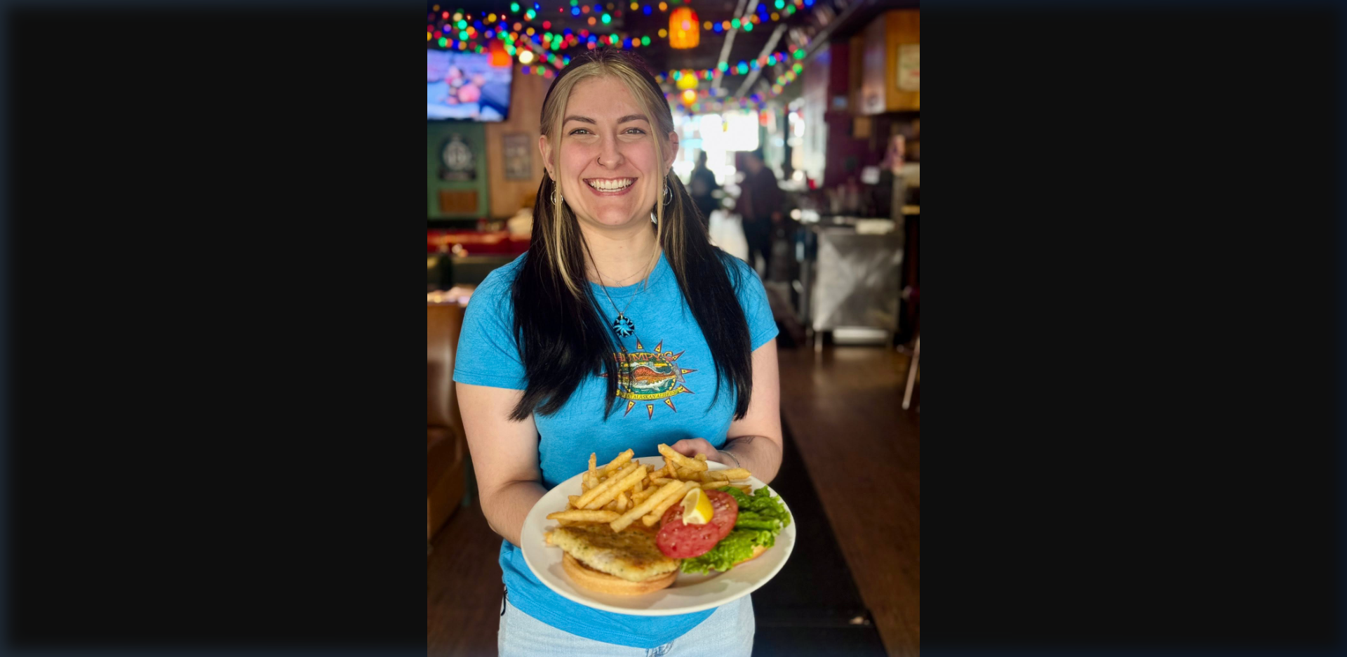 Humpy's staff member smiling and holding a plate of halibut sandwich with fries in the Humpy's Great Alaskan Alehouse dining room, wearing the Humpy's branded blue t-shirt, string lights overhead