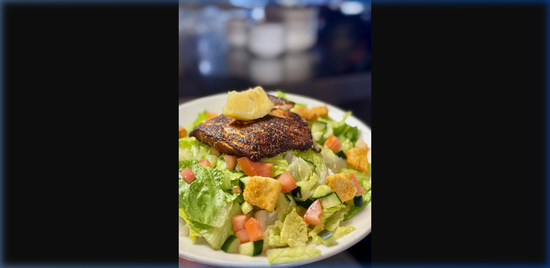 Blackened Alaska fish fillet with Cajun seasoning on a bed of crisp romaine salad with tomatoes, cucumber, and croutons, topped with a lemon wedge — Humpy's bar atmosphere in background