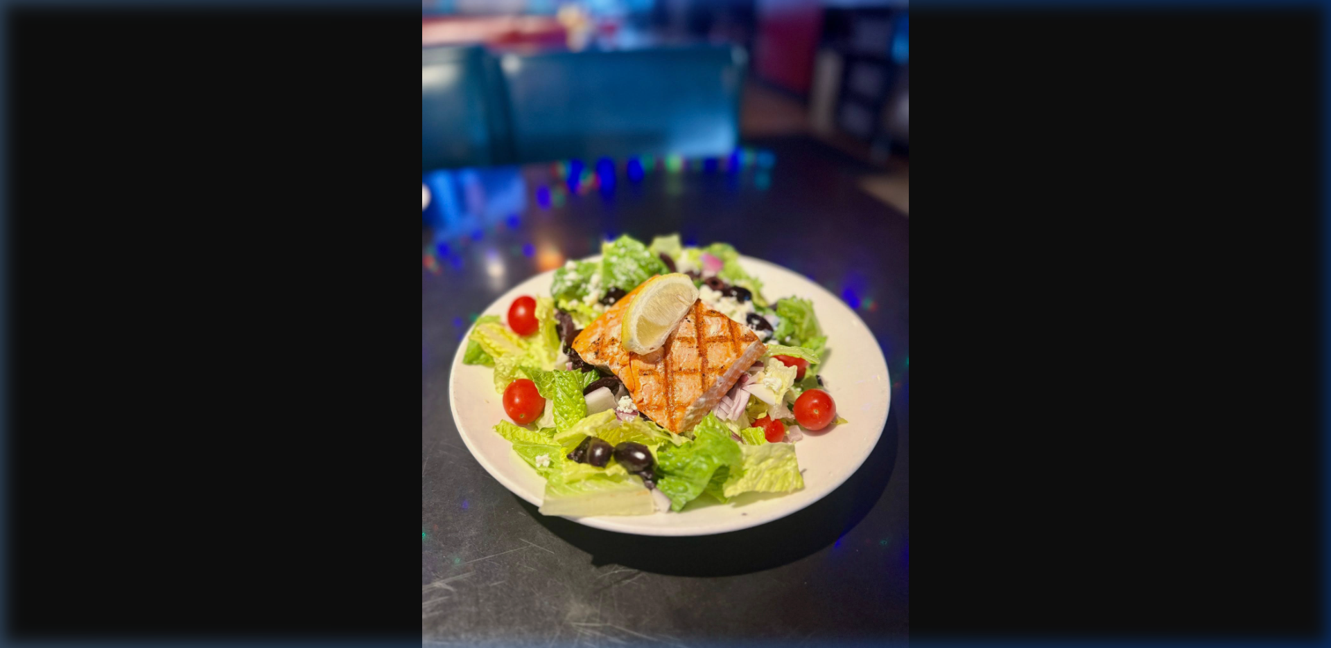 Grilled wild Alaska salmon fillet with cross-hatch grill marks on a fresh garden salad with cherry tomatoes, olives, and lemon — bar atmosphere visible in background at Humpy's
