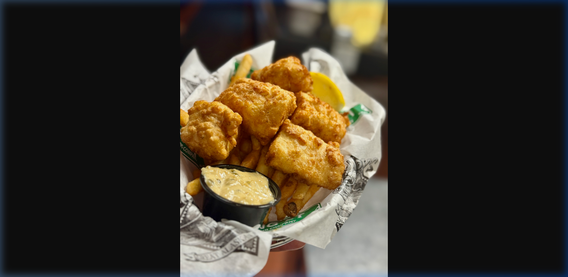 Golden beer-battered Alaska halibut fish and chips in a basket lined with Alaskan Brewing Company paper, served with fries, tartar sauce, and lemon at Humpy's Alehouse