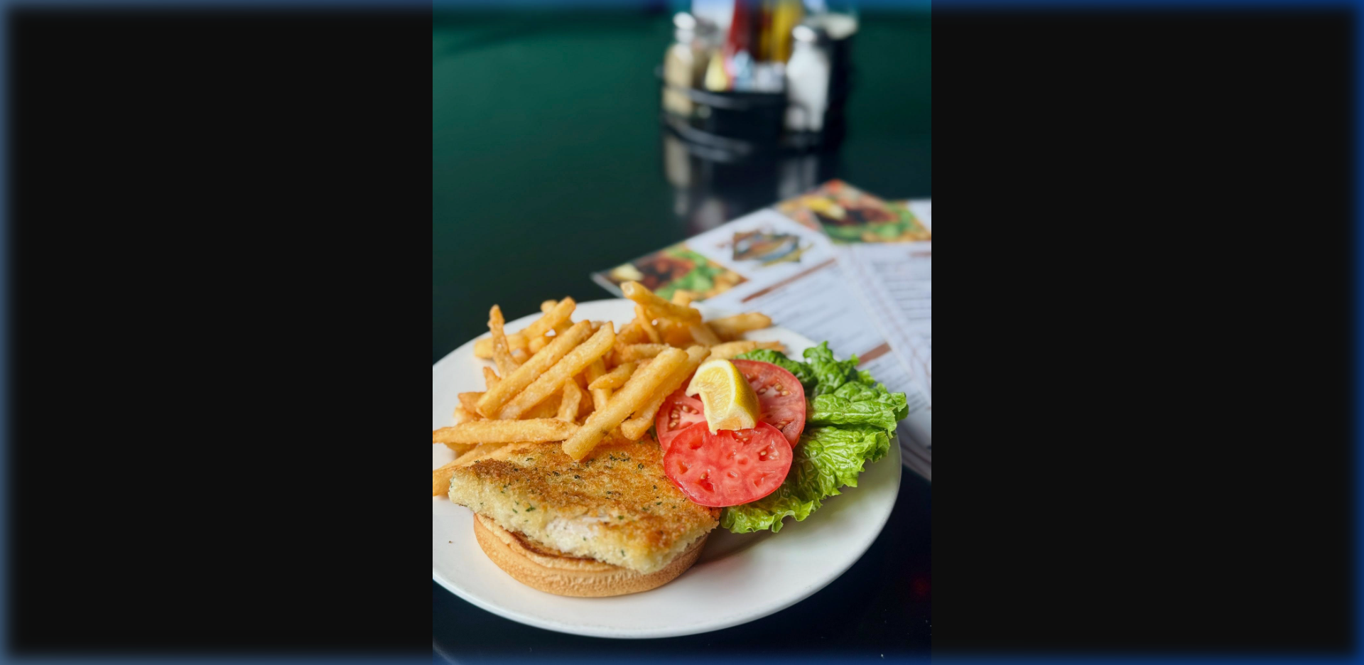 Close-up of a golden pan-fried halibut sandwich with crispy fries, tomato, and lettuce on a white plate at Humpy's — Humpy's branded menu visible in background