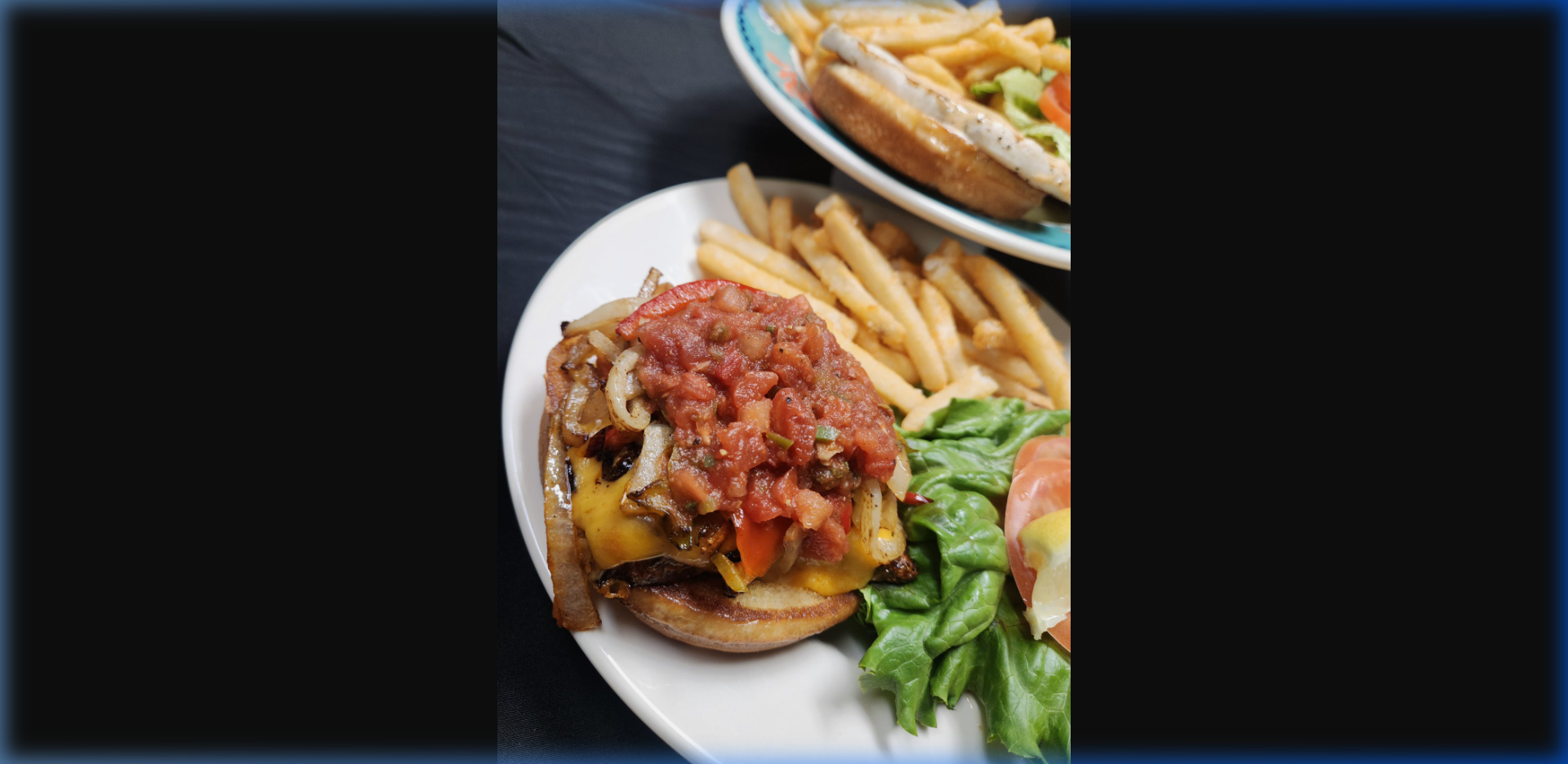 Humpy's loaded burger topped with melted cheddar, caramelized onions, and fresh salsa on a toasted bun with crispy fries — second sandwich visible in background